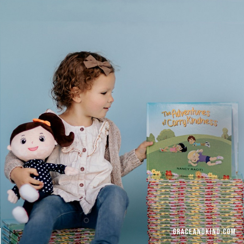 A small child holding a plush doll and holding up the Carry Kindness book