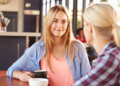 Blonde woman sitting across from another women at a cafe with a cup of coffee in her hand