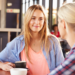 Blonde woman sitting across from another women at a cafe with a cup of coffee in her hand